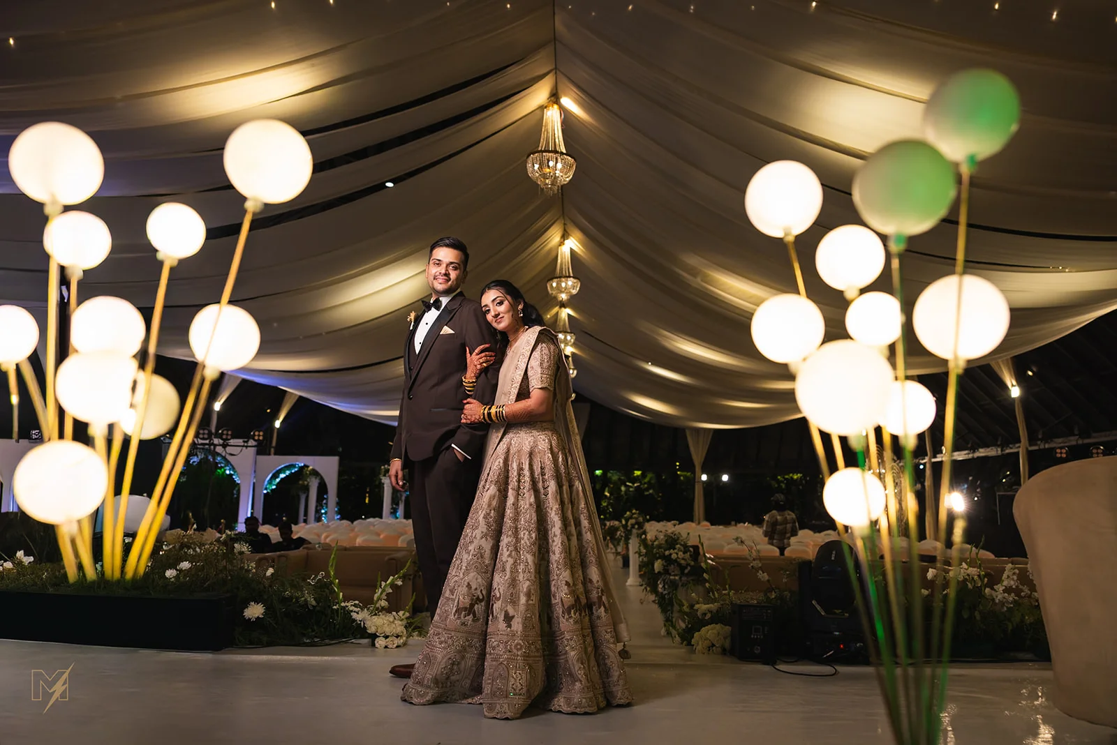 Indian bride and groom posing under illuminated ceiling draping surrounded by warm globe lights at their wedding reception.