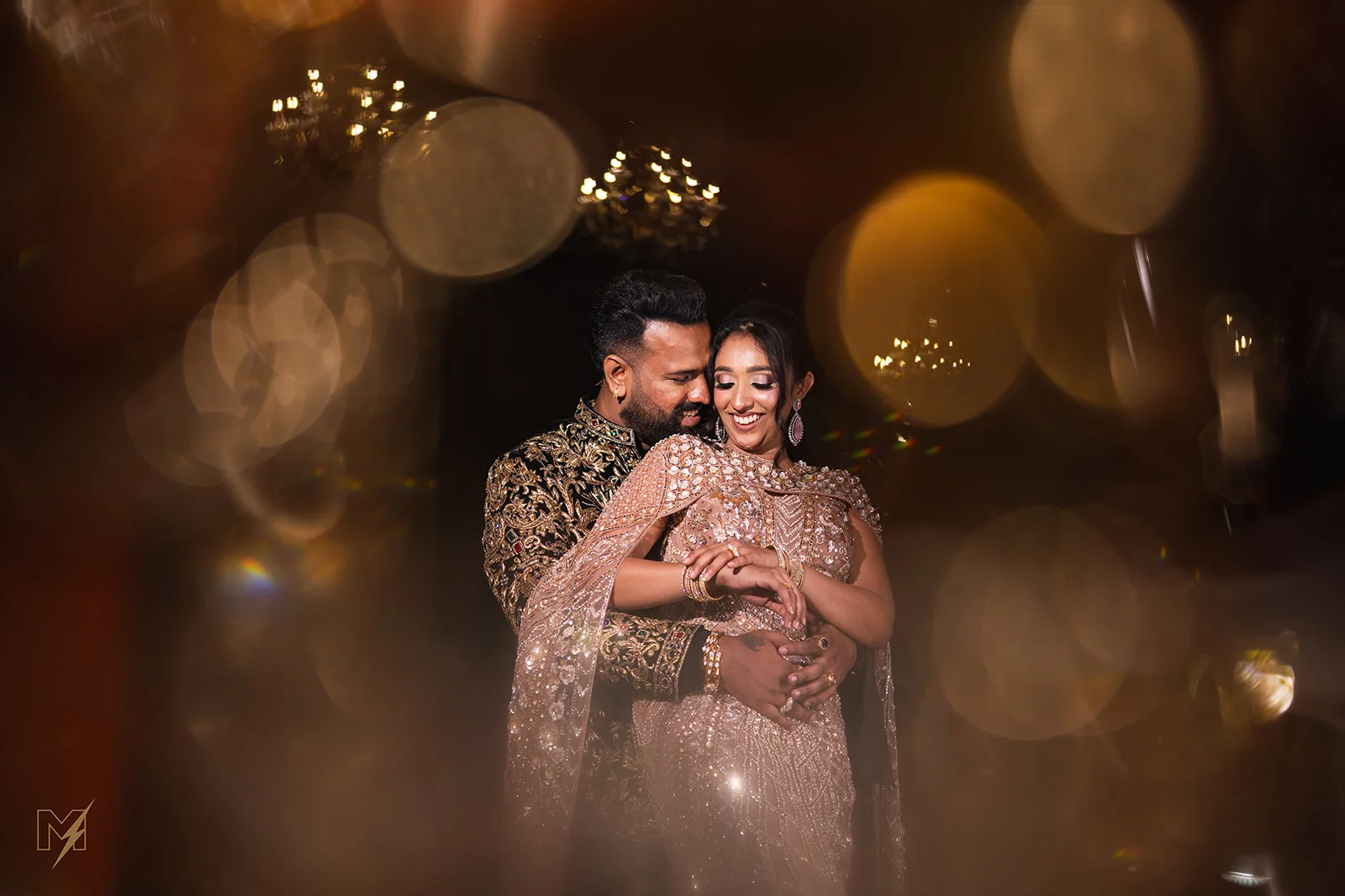 Groom embracing smiling bride in embellished outfit surrounded by warm golden bokeh light foreground.