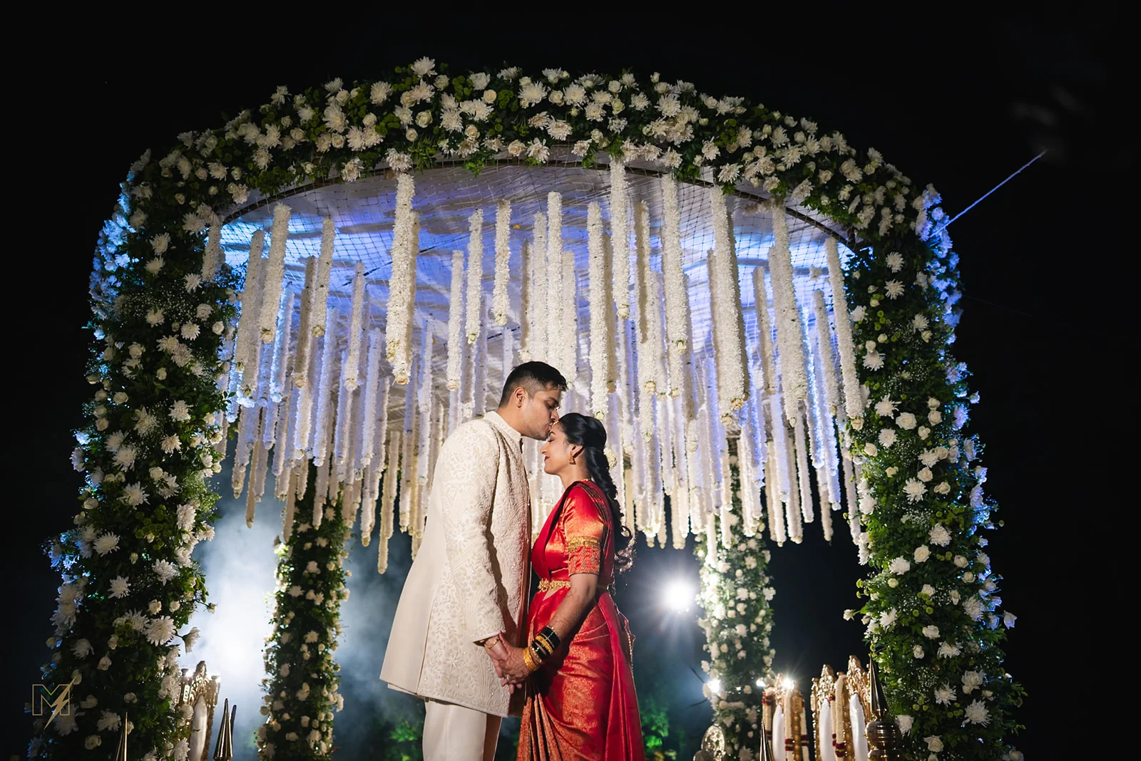 Groom kissing bride's forehead under a white floral mandap at night