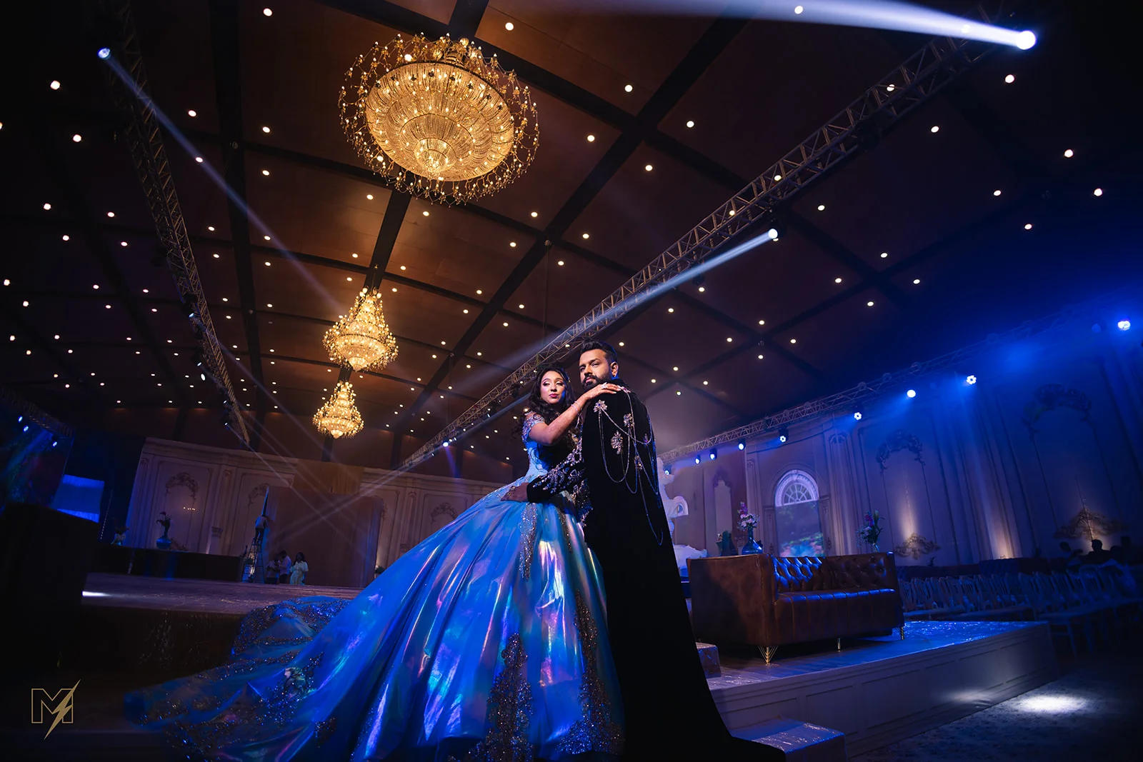 An elegant couple embracing under blue spotlights and chandeliers at a grand wedding reception.